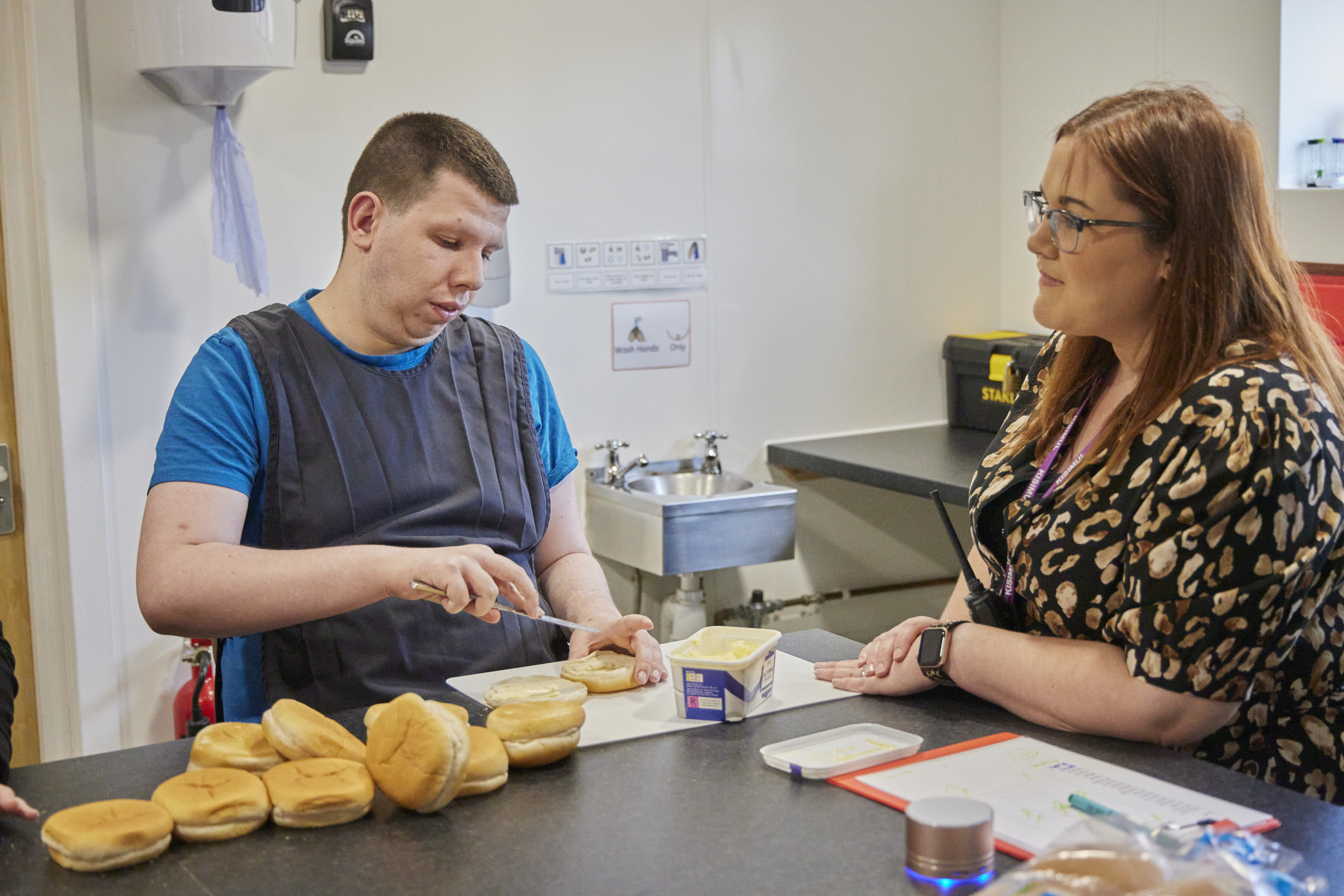 Man spreading butter on buns with a woman watching. Kitchen setting.